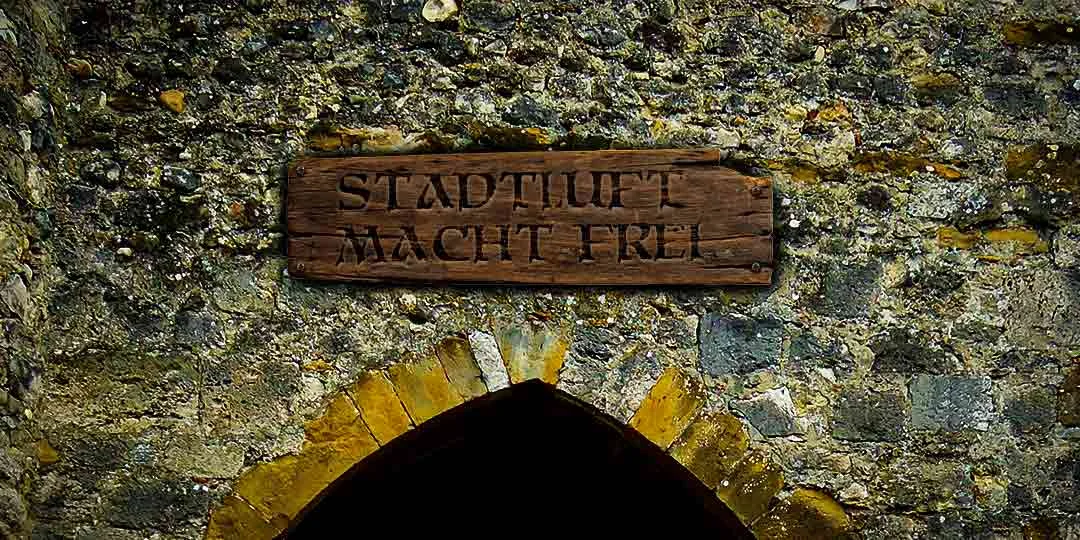 A weathered stone wall with an arched opening at the bottom, above which is affixed a wooden sign reading "STADTLUFT MACHT FREI" in carved capital letters [gen AI description].